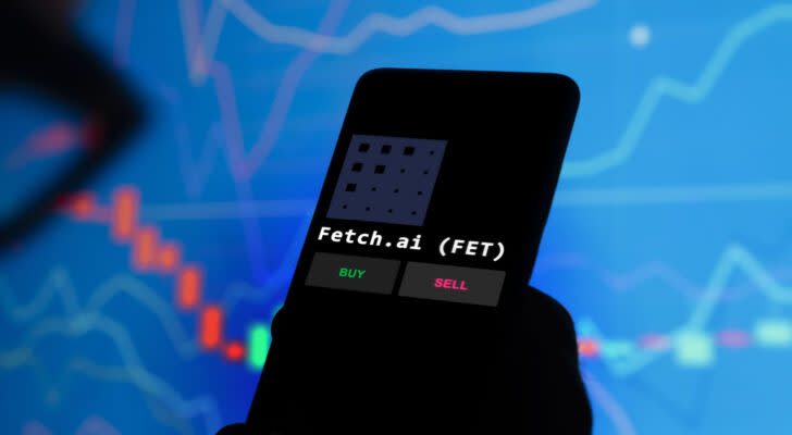 Fetch.AI (FET) cryptographic logo displayed on a smartphone with words "buy" And "sell" below the logo and the financial table in the background